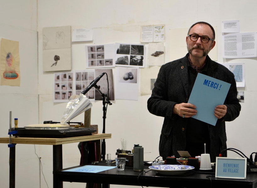 Thierry Weyd, devant ses différents objets et dispositifs sonores, et tenant un papier sur lequel est écrit 